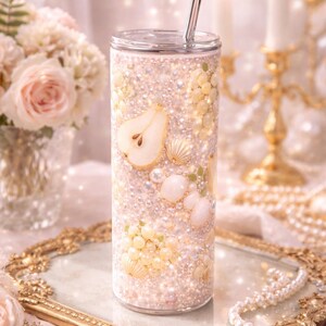 Puede incluir: Un vaso cilíndrico con tapa transparente y pajita de metal, con un exterior decorativo cubierto de pequeñas perlas blancas, cuentas color crema, conchas pequeñas y un gráfico de una rodaja de pera en tonos pastel suaves.
