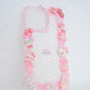 Op de afbeelding: Transparante telefoonhoes met een roze rand en decoratieve roze versieringen. De hoes is versierd met verschillende schattige, cartoonachtige figuren, strikken en aardbeien. De hoes heeft een glitterinterieur.