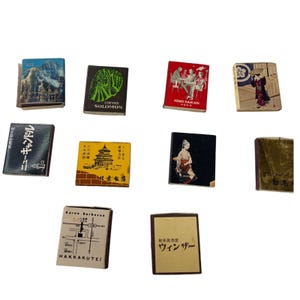 May include: A collection of ten rectangular matchboxes featuring various graphic designs, including illustrations of landscapes, figures in traditional clothing, architectural structures, maps, and Japanese text printed on the covers.