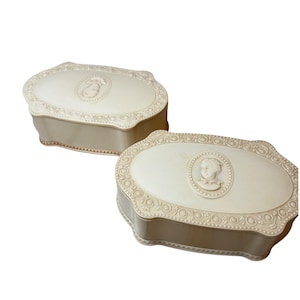 May include: Two off-white, oval-shaped decorative boxes. Each box features a raised relief portrait on the lid, surrounded by ornate scrollwork. The boxes have a scalloped edge and a neutral color scheme.