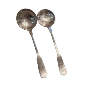 May include: Two antique silver ladles with round bowls and long, slender handles. The handles have a decorative, engraved design near the base. The ladles are displayed against a white background, highlighting their reflective surfaces.