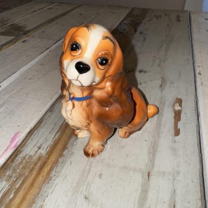 May include: A ceramic figurine of a dog, featuring brown and white fur with a blue collar. The dog is sitting, with a detailed face and large, expressive eyes. The figurine is on a weathered, white wooden surface.