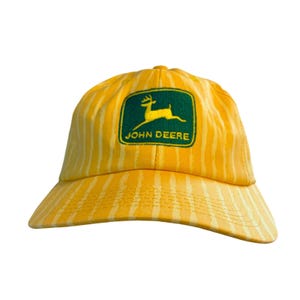 May include: Yellow and white striped baseball cap with the John Deere logo. The logo features a green rectangle with a yellow deer and the words "JOHN DEERE" in white. The cap has a curved brim.