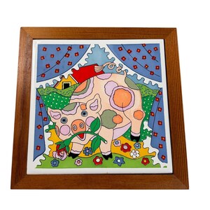 May include: A square ceramic tile framed in wood featuring a colorful illustration of a pig with pink, orange, and green spots holding a pink rose in its mouth. The background depicts a green field, a red and yellow house, and blue curtains.