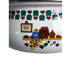 May include: White enamelware pot with a decorative apple pie design. The design includes a sign that says "Homemade Apple Pie", a basket of apples, and a jar of apple jam. The rim of the pot has a pattern of green and red shapes.
