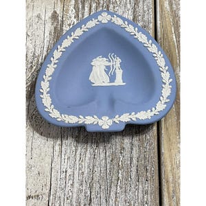 May include: A heart-shaped, light blue dish with a white floral border and a central relief design. The dish features a classical scene in white relief. The dish is likely made of ceramic or porcelain.
