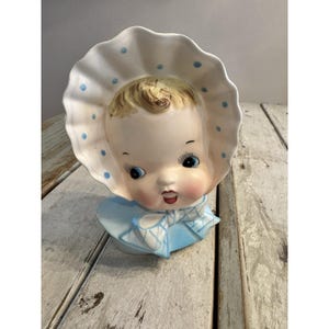 May include: A vintage ceramic baby head vase. The baby has blue eyes, rosy cheeks, and a smiling expression. The baby is wearing a white bonnet with blue polka dots and a blue bow. The vase is light blue and heart-shaped.