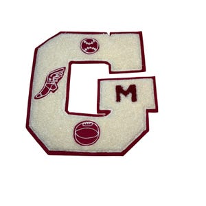 May include: A white, fuzzy letter "G" patch with a maroon border. The patch features a baseball, a track shoe with wings, and a basketball. The letter "M" is also visible on the patch.