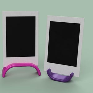 May include: Two blank polaroid-style photo frames with black centers and white borders. One frame is propped up by a bright pink stand, the other by a purple stand. The background is a light green color.