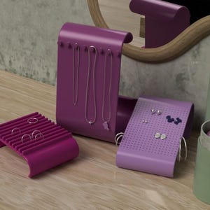 May include: A set of jewellery organisers in shades of purple and lavender. The set includes a ring holder, a necklace display, and an earring holder. The organisers are on a wooden surface, with a green container and a mirror in the background.