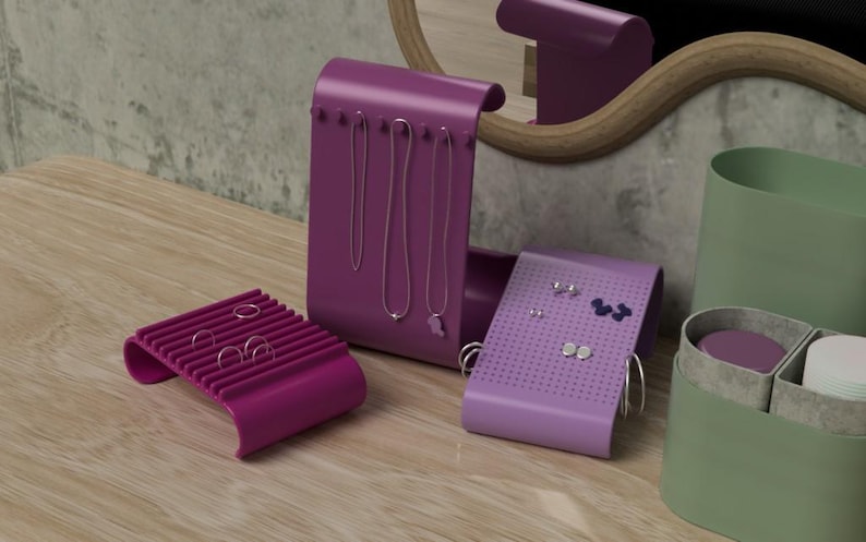 May include: A collection of purple and lavender jewelry organizers. The set includes a ring holder, a necklace display, and an earring holder. The organizers are on a wooden surface, with a green container and a mirror in the background.