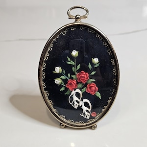 May include: An oval-shaped gold-toned metal frame featuring a black background. Inside the frame is a decorative arrangement of red and white flowers with green leaves, positioned above two white seashells. The frame has a small top loop.