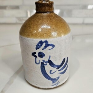 May include: A small stoneware jug with a cylindrical body and a narrow neck. The lower portion is off-white with a hand-painted blue rooster design, while the upper neck and rim are finished in a glossy, speckled brown glaze.