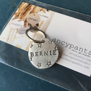 K&ouml;nnte beinhalten: Ein runder Schl&uuml;sselanh&auml;nger aus geb&uuml;rstetem, silberfarbenem Metall mit dem mittig eingestanzten Namen BERNIE in Gro&szlig;buchstaben, umgeben von kleinen eingravierten Sternmotiven. Der Anh&auml;nger ist an einem runden Metallring befestigt.