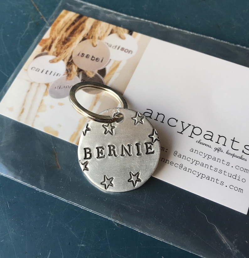 K&ouml;nnte beinhalten: Ein runder Schl&uuml;sselanh&auml;nger aus geb&uuml;rstetem, silberfarbenem Metall mit dem mittig eingestanzten Namen BERNIE in Gro&szlig;buchstaben, umgeben von kleinen eingravierten Sternmotiven. Der Anh&auml;nger ist an einem runden Metallring befestigt.