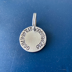 K&ouml;nnte beinhalten: Eine kreisf&ouml;rmige Haustiermarke aus silberfarbenem Metall mit geb&uuml;rsteter Oberfl&auml;che. Der Rand der Marke ist mit den Zahlen 01234567890 in einem kreisf&ouml;rmigen Muster gepr&auml;gt. Ein kleiner silberfarbener Spaltring ist oben an der Marke befestigt.