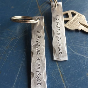 May include: Two silver keychains with engraved coordinates. The top keychain has the coordinates 37°47' 12" N 122° 24' 2" W. The bottom keychain has the coordinates 38°3' 44" N 78°43′40″W.