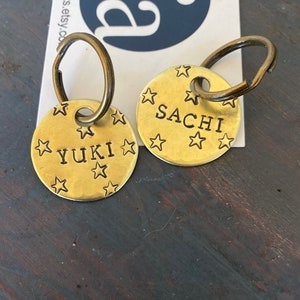 K&ouml;nnte beinhalten: Zwei runde Haustiermarken aus Messing mit dunklen Metallringen. Jede Marke ist mit eingestanzten Sternmustern sowie den Namen YUKI und SACHI in schwarzer Schrift versehen und liegt auf einer wei&szlig;en Karte mit blauem Logo und dem Text ancypants.etsy.com.