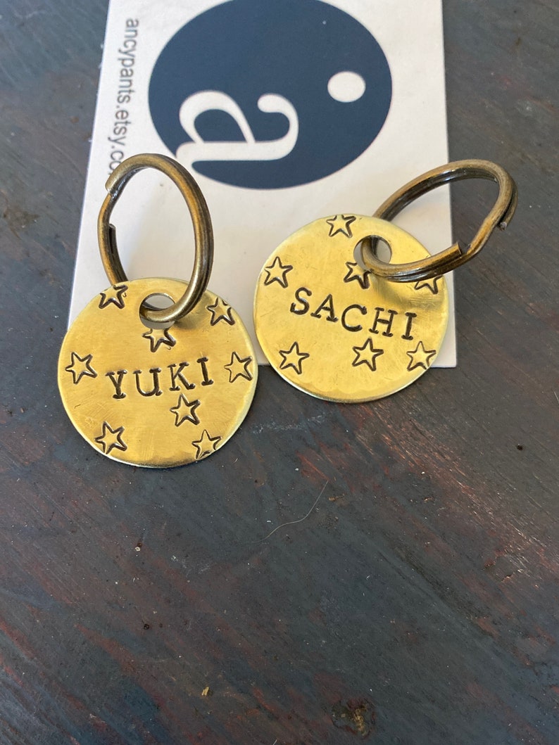 K&ouml;nnte beinhalten: Zwei runde Haustiermarken aus Messing mit dunklen Metallringen. Jede Marke ist mit eingestanzten Sternmustern sowie den Namen YUKI und SACHI in schwarzer Schrift versehen und liegt auf einer wei&szlig;en Karte mit blauem Logo und dem Text ancypants.etsy.com.