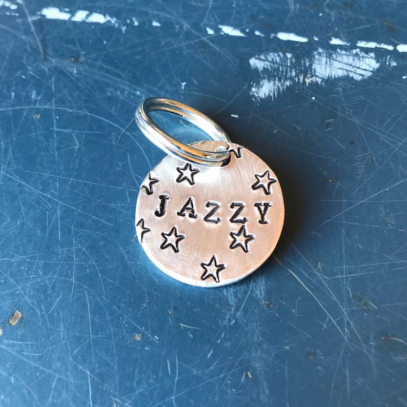 K&ouml;nnte beinhalten: Eine kreisf&ouml;rmige Haustiermarke aus silberfarbenem Metall mit dem mittig eingestanzten Namen JAZZY in Gro&szlig;buchstaben, umgeben von mehreren kleinen eingravierten Sternen und befestigt an einem silberfarbenen Metall-Spaltring.