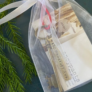 May include: A small, gold-colored skeleton key with a tag that reads "450 year old ruff AW" is inside a white organza bag with a red ribbon. The bag is on a dark green surface.