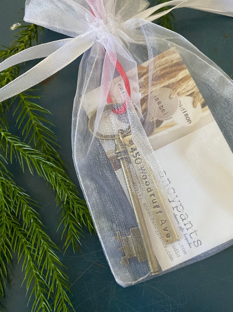 May include: A small, gold-colored skeleton key with a tag that reads "450 year old ruff AW" is inside a white organza bag with a red ribbon. The bag is on a dark green surface.