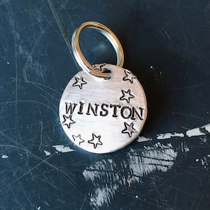 K&ouml;nnte beinhalten: Eine runde Haustiermarke aus silberfarbenem Metall mit dem eingestanzten Namen WINSTON in Gro&szlig;buchstaben in der Mitte. Die Marke ist mit kleinen eingestanzten Sternen verziert und verf&uuml;gt &uuml;ber einen runden Metallring zur Befestigung.