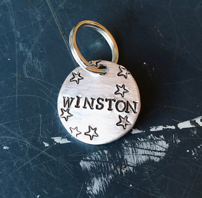 K&ouml;nnte beinhalten: Eine runde Haustiermarke aus silberfarbenem Metall mit dem eingestanzten Namen WINSTON in Gro&szlig;buchstaben in der Mitte. Die Marke ist mit kleinen eingestanzten Sternen verziert und verf&uuml;gt &uuml;ber einen runden Metallring zur Befestigung.