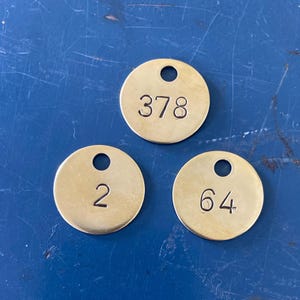 May include: Three small, round, brass tags with a hole at the top. Each tag has a different number stamped on it: 378, 2, and 64. The tags are on a blue surface.