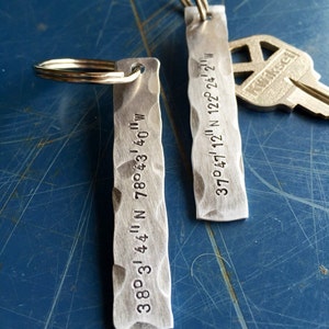 May include: Two rectangular, hammered metal keychains with stamped coordinates and key rings. One keychain reads "38° 3' 44" N 78° 43' 40" W" and the other reads "37° 47' 12" N 122° 24' 2" W". A silver key is in the background.