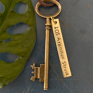 May include: A gold-toned skeleton key necklace with a brown leather cord. The key has a tag attached to it that reads "105 Arlington Street".