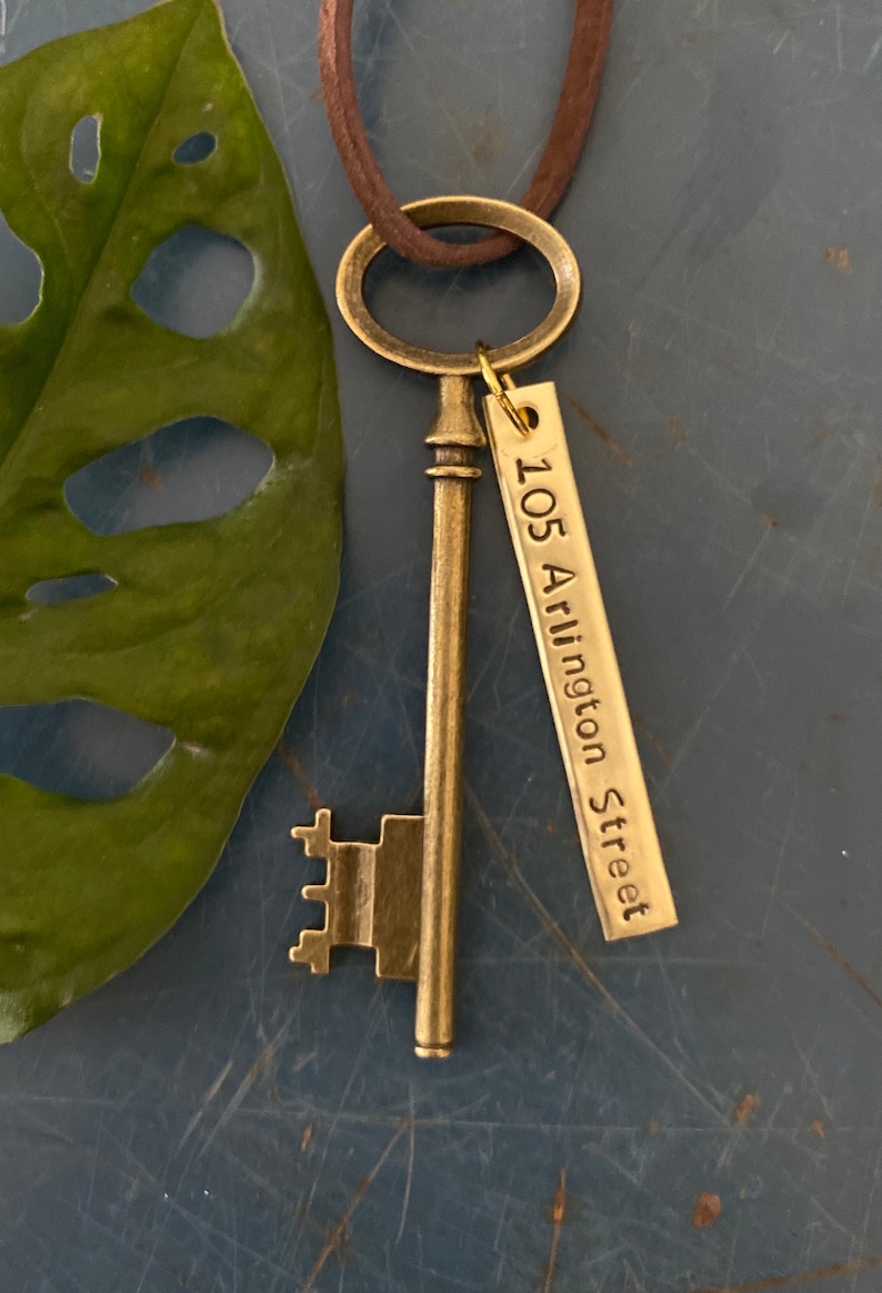 May include: A gold-toned skeleton key necklace with a brown leather cord. The key has a tag attached to it that reads "105 Arlington Street".