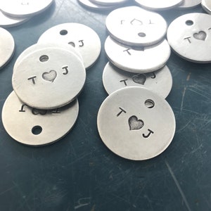 May include: A close-up of several silver metal round tags with the letters "T" and "J" and a heart symbol engraved on them. The tags have a hole in the center.