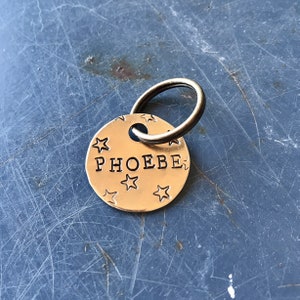 K&ouml;nnte beinhalten: Eine runde Haustiermarke aus goldfarbenem Metall mit dem eingestanzten Namen PHOEBE in Gro&szlig;buchstaben in der Mitte. Die Oberfl&auml;che der Marke ist mit mehreren kleinen, eingestanzten Sternen verziert und enth&auml;lt einen runden Metallring.