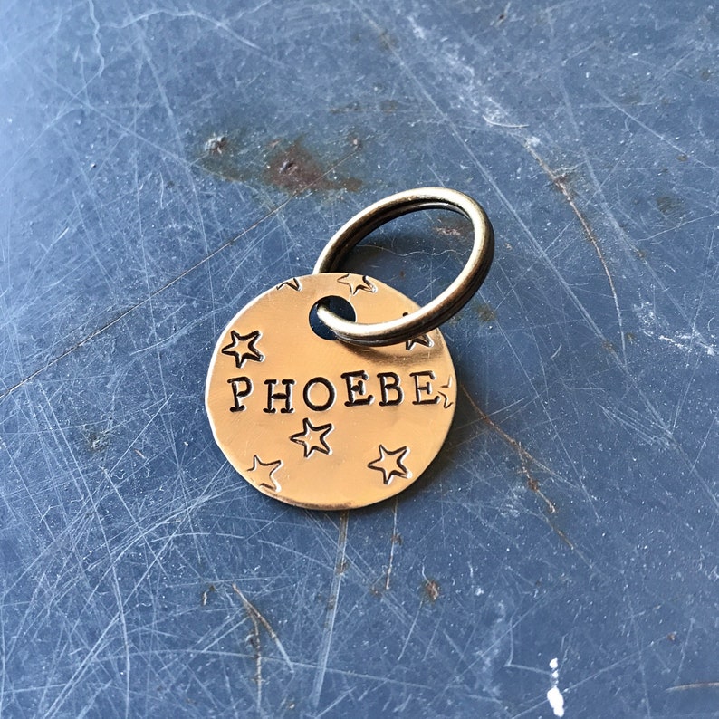 K&ouml;nnte beinhalten: Eine runde Haustiermarke aus goldfarbenem Metall mit dem eingestanzten Namen PHOEBE in Gro&szlig;buchstaben in der Mitte. Die Oberfl&auml;che der Marke ist mit mehreren kleinen, eingestanzten Sternen verziert und enth&auml;lt einen runden Metallring.