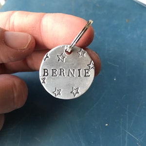 K&ouml;nnte beinhalten: Eine kreisf&ouml;rmige Haustiermarke aus silberfarbenem Metall mit dem eingestanzten Namen BERNIE in Gro&szlig;buchstaben in der Mitte, umgeben von mehreren kleinen eingestanzten Sternformen und befestigt an einem kleinen silberfarbenen Metallring.