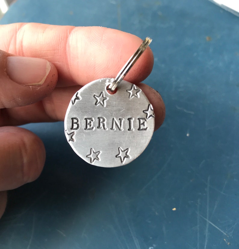 K&ouml;nnte beinhalten: Eine kreisf&ouml;rmige Haustiermarke aus silberfarbenem Metall mit dem eingestanzten Namen BERNIE in Gro&szlig;buchstaben in der Mitte, umgeben von mehreren kleinen eingestanzten Sternformen und befestigt an einem kleinen silberfarbenen Metallring.