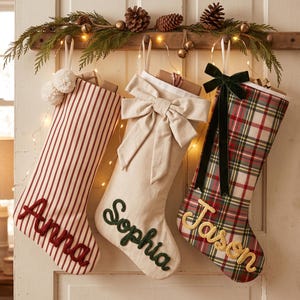 Peut inclure: Trois chaussettes de Noël suspendues avec des prénoms brodés en écriture cursive. La première est rayée rouge et blanc avec des pompons, la deuxième est crème unie avec un grand nœud, et la troisième est à carreaux rouges et verts avec un nœud en velours.