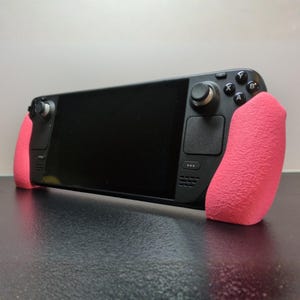 May include: A black handheld gaming console with a large screen and pink textured grips on each side. The console has buttons, a joystick, and a touchpad. The device is on a dark surface.