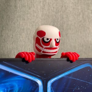 Op de afbeelding: Een wit en rood beeldje van een Titan uit de anime-serie Attack on Titan. Het beeldje heeft een gedetailleerd gezicht met rode markeringen en houdt zich met rode handen vast aan de rand van een donker oppervlak.