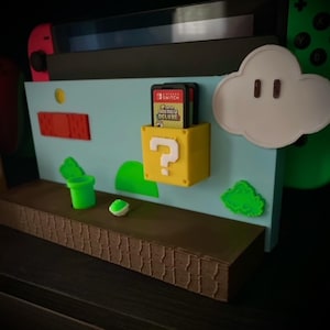 May include: A 3D-printed Nintendo Switch game cartridge holder with a Super Mario Bros. theme. The holder features a yellow question mark block, a white cloud, and other game elements. The background is light blue with brown and green accents.