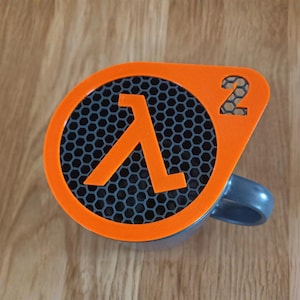 Peut inclure: Une tasse grise avec un couvercle orange représentant le logo Half-Life 2. Le couvercle affiche le symbole lambda et le chiffre 2. La tasse est posée sur une surface en bois.