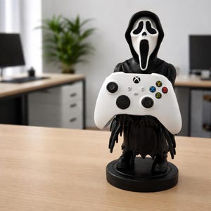 May include: A black and white Ghostface figure holding a white Xbox controller. The figure has a black robe and a round black base. The controller has black buttons and colored action buttons.