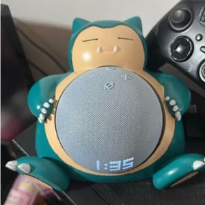 Op de afbeelding: Een teal en tan Snorlax-vormige slimme luidspreker met een grijs luidsprekermesh. De tijd 1:35 wordt weergegeven in blauwe digitale cijfers op de luidspreker. De luidspreker heeft een ronde buik en zit.