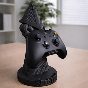 Könnte beinhalten: Ein schwarzer Videospiel-Controller auf einem schwarzen Ständer mit Pyramid Head-Design. Der Ständer trägt die Aufschrift "Silent Hill". Der Controller hat farbige Tasten und ein Steuerkreuz.