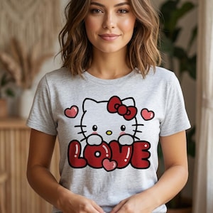 May include: A light gray t-shirt featuring a graphic of Hello Kitty holding the word "LOVE" in red bubble letters. The design includes a red bow, hearts, and black outlines. The shirt has short sleeves.