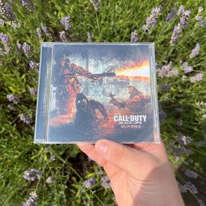 Könnte beinhalten: Eine transparente CD-Hülle mit dem Spiel "Call of Duty: Black Ops Zombies". Das Cover zeigt Zombie-Figuren und einen Soldaten, der eine Waffe abfeuert, vor einem feurigen Hintergrund. Die Hülle wird vor Lavendelblüten gehalten.