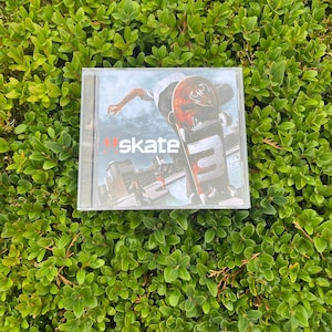 May include: A CD case with the word "skate" on the cover. The artwork shows a skateboarder performing a trick, with a city visible in the background. The case is placed on a bed of green leaves.