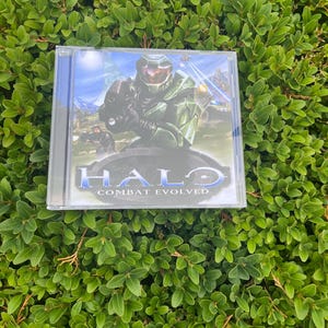 Puede incluir: Una funda de plástico transparente que contiene el videojuego "Halo: Combat Evolved". La portada del juego presenta una figura verde blindada sosteniendo un arma, con el título del juego en letras azules y plateadas. El fondo muestra un paisaje con montañas y cielo.