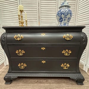 Puede incluir: Cómoda negra con tres cajones y herrajes dorados. La cómoda tiene un frente curvo y tallas decorativas. Un jarrón de porcelana azul y blanco y un candelabro dorado están encima. La cómoda mide aproximadamente 100 cm de ancho.